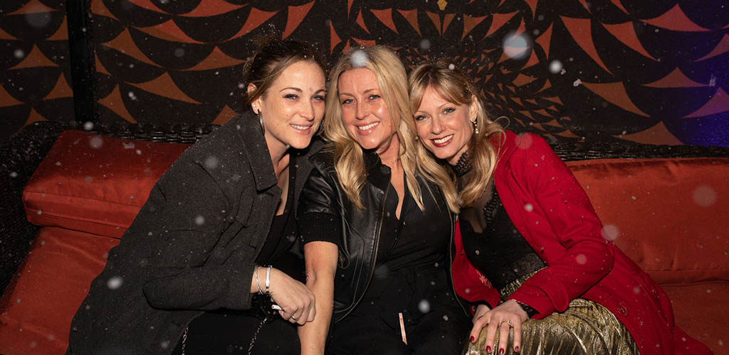 Three beautiful young woman at The Righteous Room sitting close together, smiilng, on a red couch with a red and black background and snow falling in foreground.