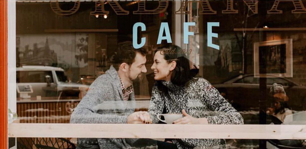 A young couple laughing intimately in a cafe window with the words CAFE over their heads