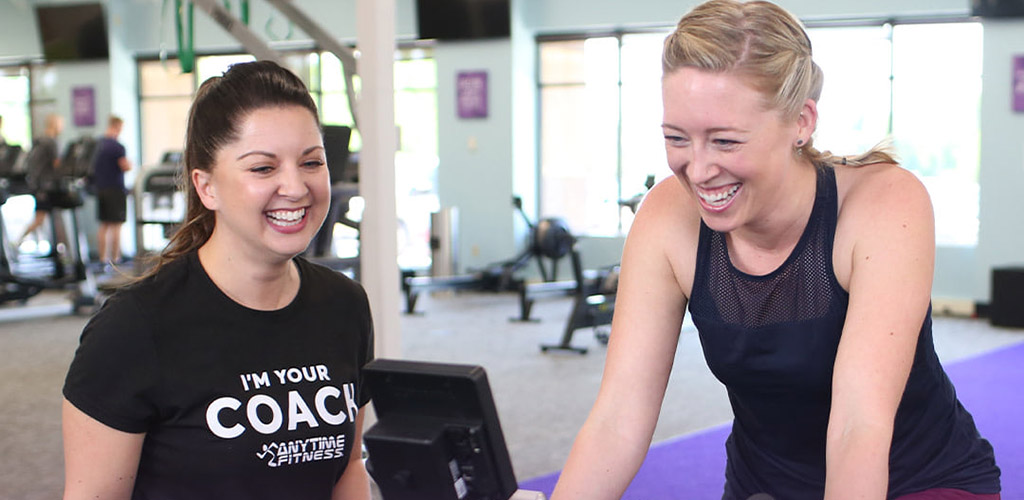 Beautiful blonde training on the stationary bike at Anytime Fitness with her petite brunette coach